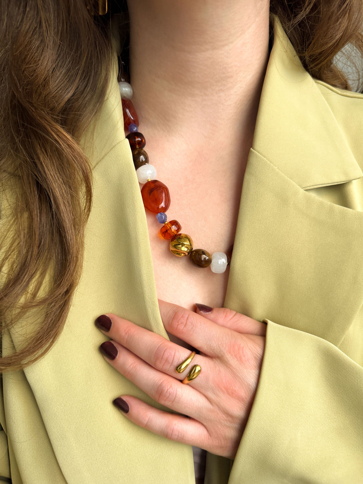 Vrouw draagt een kralenketting met bruine, terracotta, paarse en witte kralen en een gouden organische ring uit de Atelier Colore collectie van Don’ aparte.