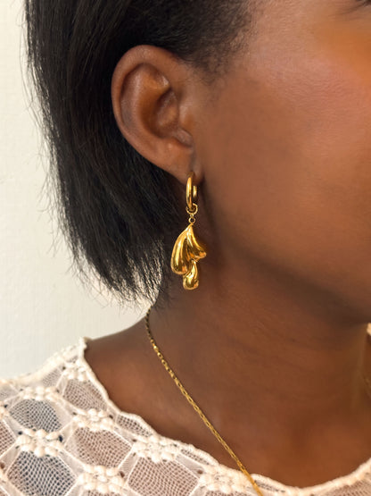 Close-up van een vrouw met gouden oorringen en een organisch gevormde druppelhanger uit de Atelier Colore collectie van Don’ aparte, gecombineerd met een witte kanten top.