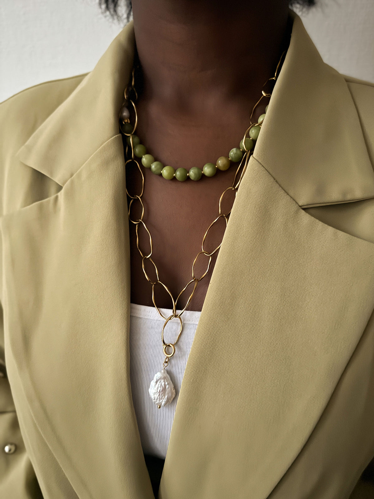 Vrouw draagt een gouden statement schakelketting met barokparel en een groene kralenketting, gecombineerd met een lichtgroene blazer.
