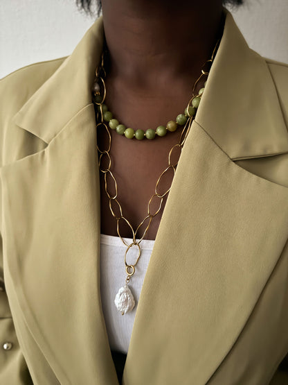 Vrouw draagt een gouden statement schakelketting met barokparel en een groene kralenketting, gecombineerd met een lichtgroene blazer.