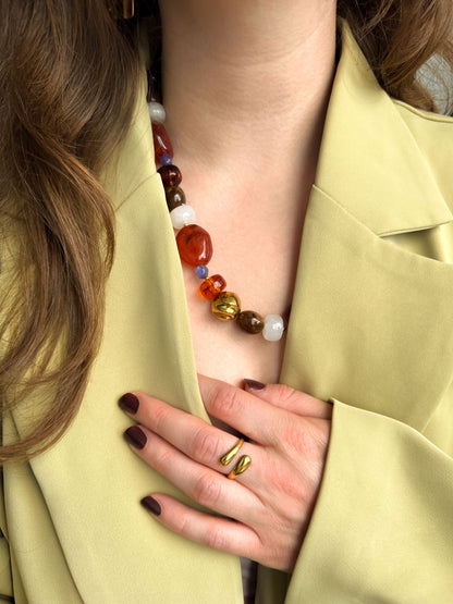 Vrouw draagt een kralenketting met bruine, terracotta, paarse en witte kralen en een gouden organische ring uit de Atelier Colore collectie van Don’ aparte.